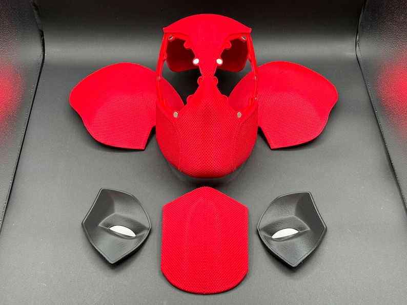 May include: A red and black 3D printed Deadpool mask with separate pieces for the eyes, mouth, and head. The mask is made of a textured material.