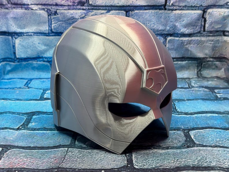 May include: A silver and red superhero helmet with a stylized design. The helmet features a smooth, metallic finish with a prominent crest on the forehead and eye openings. The color scheme is primarily silver with red accents.