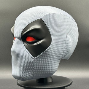 Może przedstawiać: Biała maska Deadpoola z czarną maską na oczy i czerwonymi oczami. Maska wykonana jest z fakturowanego materiału i stoi na czarnym stojaku.