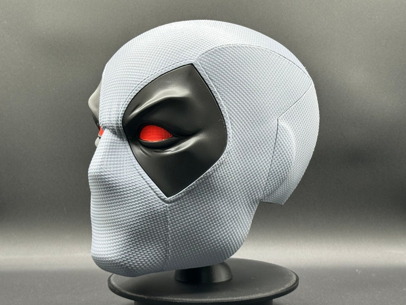 Może przedstawiać: Biała maska Deadpoola z czarną maską na oczy i czerwonymi oczami. Maska wykonana jest z fakturowanego materiału i stoi na czarnym stojaku.