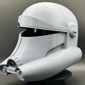 May include: A gray 3D printed helmet with a raised design. The helmet has a hinged visor and a detailed design on the front.