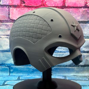 May include: A gray helmet with a star emblem and a woven pattern on the side. The helmet has eye openings and is displayed on a black stand. The background is a brick wall with blue, pink, and red colors.