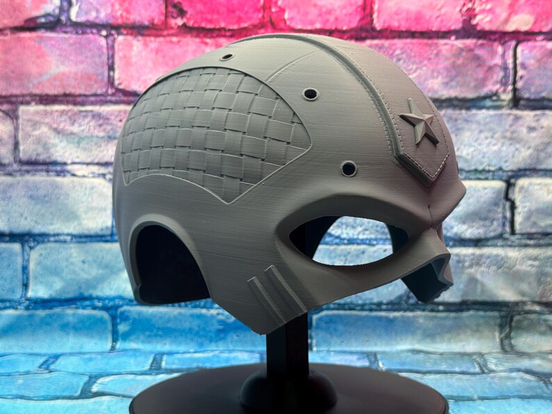 May include: A gray helmet with a star emblem and a woven pattern on the side. The helmet has eye openings and is displayed on a black stand. The background is a brick wall with blue, pink, and red colors.