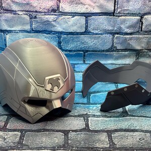 May include: A silver-colored helmet and face mask set. The helmet has a stylized design with a bird emblem. The face mask is a separate piece, dark gray in color, with a geometric shape and rivet details.