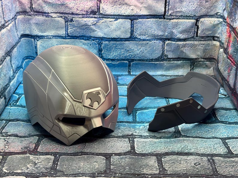 May include: A silver-colored helmet and face mask set. The helmet has a stylized design with a bird emblem. The face mask is a separate piece, dark gray in color, with a geometric shape and rivet details.