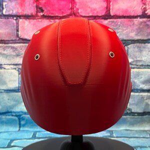 May include: A vibrant red helmet with a smooth, rounded design. The helmet features a series of small, silver-colored rivets and a central seam detail. The helmet is displayed on a black stand against a brick wall background.