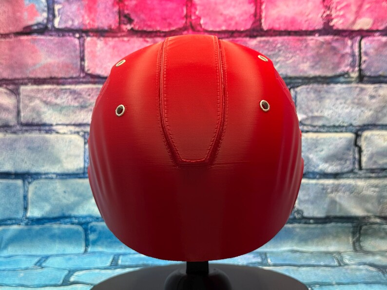 May include: A vibrant red helmet with a smooth, rounded design. The helmet features a series of small, silver-colored rivets and a central seam detail. The helmet is displayed on a black stand against a brick wall background.