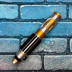 May include: A silver and gold lightsaber prop with black accents. The hilt features a black grip section with vertical lines, and the blade emitter has a gold finish with small holes. The lightsaber is set against a blue brick background.