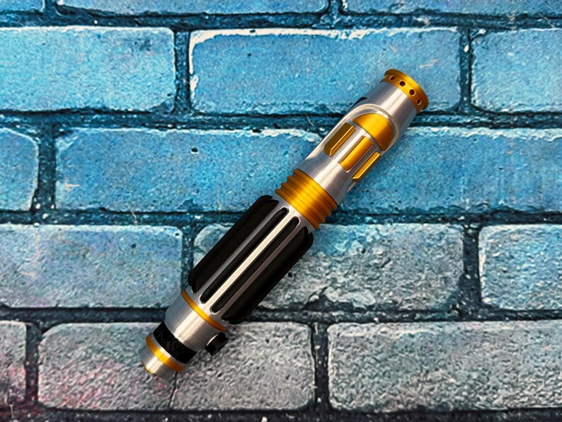 May include: A silver and gold lightsaber prop with black accents. The hilt features a black grip section with vertical lines, and the blade emitter has a gold finish with small holes. The lightsaber is set against a blue brick background.