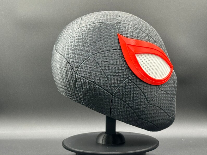 Puede incluir: Una m&aacute;scara de Spider-Man impresa en 3D de color negro y rojo con un dise&ntilde;o de telara&ntilde;a texturizado. La m&aacute;scara tiene una pieza de ojo de malla blanca con un contorno rojo.
