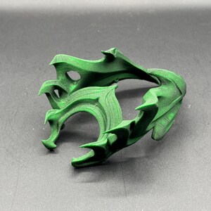 Puede incluir: Una escultura verde abstracta impresa en 3D con un dise&ntilde;o complejo y fluido. El objeto tiene bordes afilados y curvos y una superficie texturizada. La escultura se muestra sobre una superficie gris.