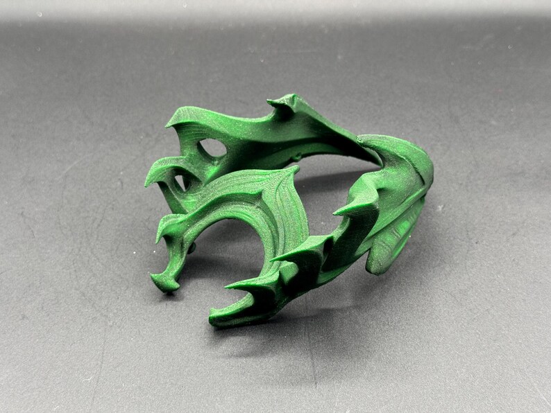 Puede incluir: Una escultura verde abstracta impresa en 3D con un dise&ntilde;o complejo y fluido. El objeto tiene bordes afilados y curvos y una superficie texturizada. La escultura se muestra sobre una superficie gris.