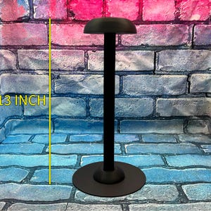 May include: A black helmet stand with a round base and a curved top. The stand is 13 inches tall. The stand is designed to hold a helmet. The background is a brick wall.