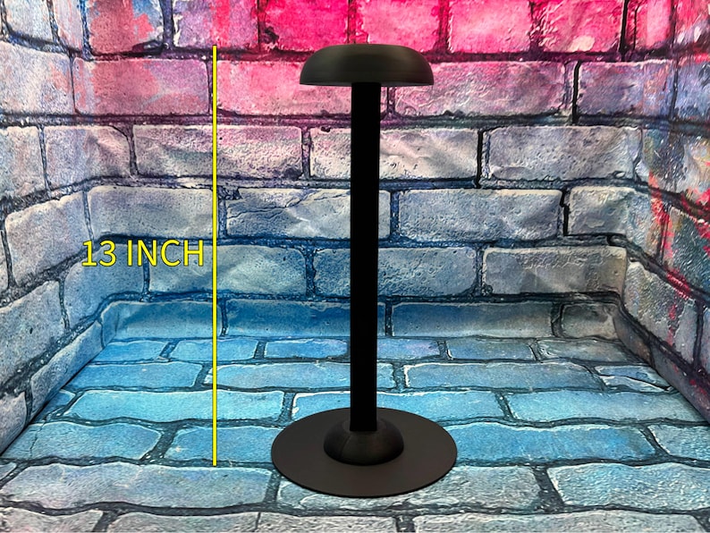 May include: A black helmet stand with a round base and a curved top. The stand is 13 inches tall. The stand is designed to hold a helmet. The background is a brick wall.