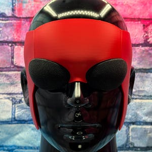 May include: A red mask with a smooth matte finish featuring two large, oval-shaped black mesh eye coverings. The mask is fitted onto a glossy black mannequin head, covering the forehead and extending down the sides to the ear area.