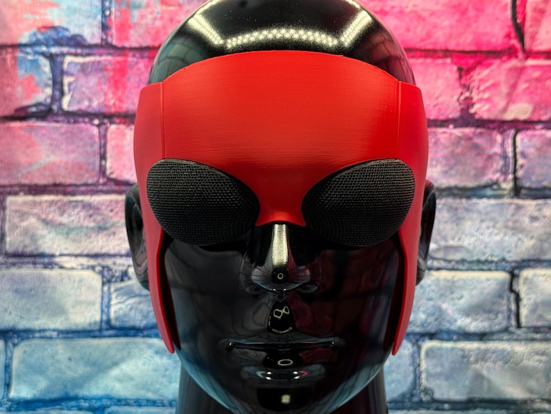 May include: A red mask with a smooth matte finish featuring two large, oval-shaped black mesh eye coverings. The mask is fitted onto a glossy black mannequin head, covering the forehead and extending down the sides to the ear area.