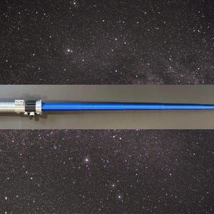 Op de afbeelding: Een blauw en zilver lichtzwaard rekwisiet, met een zwart handvatgedeelte en een helder blauw blad. Het handvat heeft gedetailleerde zilveren accenten en knoppen. De achtergrond is een sterrenhemel.
