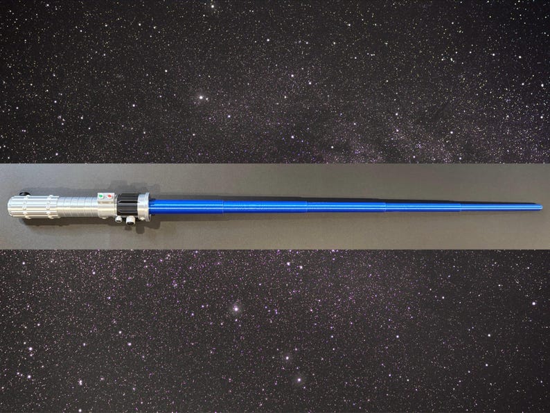 Op de afbeelding: Een blauw en zilver lichtzwaard rekwisiet, met een zwart handvatgedeelte en een helder blauw blad. Het handvat heeft gedetailleerde zilveren accenten en knoppen. De achtergrond is een sterrenhemel.