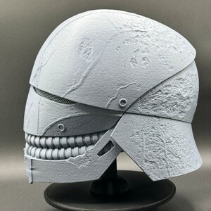 May include: A white, textured helmet with a detailed, toothy mouth design. The helmet is made of a hard plastic material and has a distressed, weathered look.