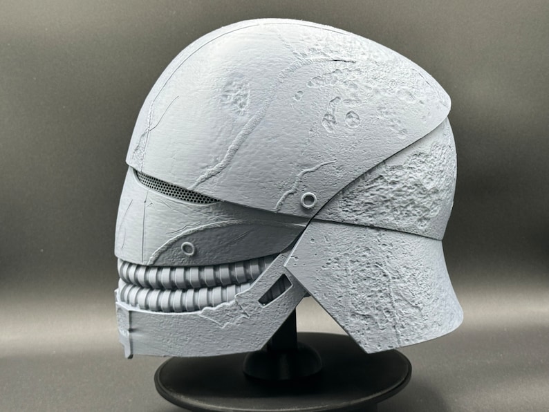 May include: A white, textured helmet with a detailed, toothy mouth design. The helmet is made of a hard plastic material and has a distressed, weathered look.