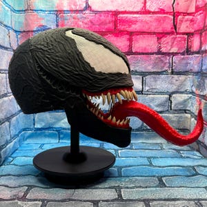 May include: A black Venom mask with a white mesh eye area, sharp white teeth, and a long red tongue. The mask is displayed on a black stand against a brick wall backdrop. The mask is a replica of the Marvel Comics character.