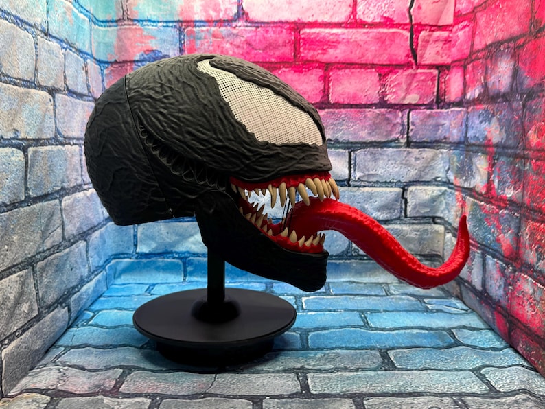 May include: A black Venom mask with a white mesh eye area, sharp white teeth, and a long red tongue. The mask is displayed on a black stand against a brick wall backdrop. The mask is a replica of the Marvel Comics character.