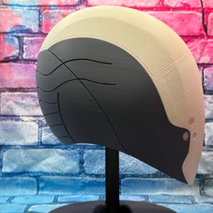 May include: A futuristic-looking helmet in shades of gray and beige, mounted on a black stand. The helmet has a smooth, curved design with textured and patterned sections. The background features a brick wall with blue, pink, and purple hues.