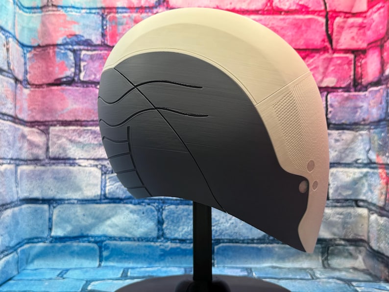 May include: A futuristic-looking helmet in shades of gray and beige, mounted on a black stand. The helmet has a smooth, curved design with textured and patterned sections. The background features a brick wall with blue, pink, and purple hues.