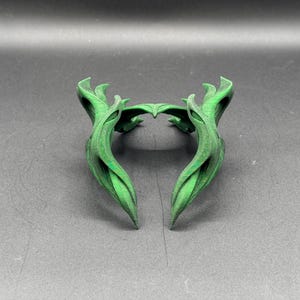 Puede incluir: Un tocado decorativo verde y estilizado con un dise&ntilde;o en forma de hoja. El tocado est&aacute; hecho de un material s&oacute;lido y tiene una forma sim&eacute;trica con extremos puntiagudos. El objeto est&aacute; sobre un fondo gris oscuro.