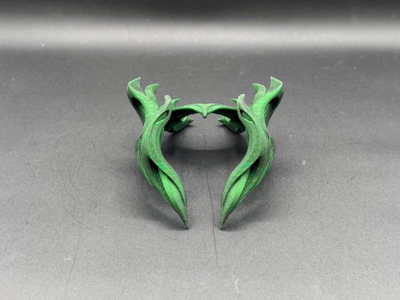 Puede incluir: Un tocado decorativo verde y estilizado con un dise&ntilde;o en forma de hoja. El tocado est&aacute; hecho de un material s&oacute;lido y tiene una forma sim&eacute;trica con extremos puntiagudos. El objeto est&aacute; sobre un fondo gris oscuro.