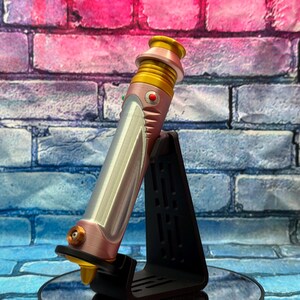 Op de afbeelding: Een roze en zilveren lichtzwaard prop met gouden accenten, rustend op een zwarte standaard. Het lichtzwaard heeft een gedetailleerd ontwerp met een rode knop en een gouden emitter. De achtergrond is een bakstenen muur met roze en blauwe tinten.