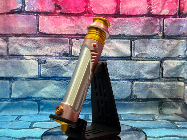 Op de afbeelding: Een roze en zilveren lichtzwaard prop met gouden accenten, rustend op een zwarte standaard. Het lichtzwaard heeft een gedetailleerd ontwerp met een rode knop en een gouden emitter. De achtergrond is een bakstenen muur met roze en blauwe tinten.