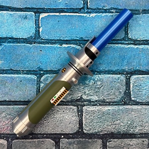 May include: A detailed lightsaber prop with a blue blade and metallic silver and green handle. The handle has intricate details and a small control panel. The lightsaber is set against a blue brick wall background.