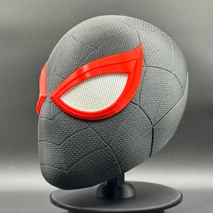 Puede incluir: Una m&aacute;scara de Spider-Man impresa en 3D de color negro y rojo con un patr&oacute;n de telara&ntilde;a y lentes de ojos rojos.