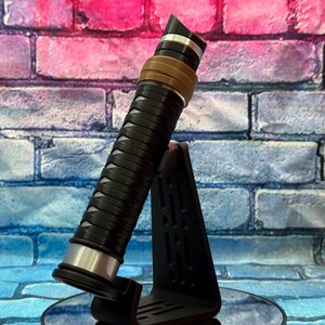 May include: A black and silver lightsaber with a brown accent ring, displayed on a black stand. The lightsaber has a textured grip and a silver hilt. The background is a colorful brick wall.