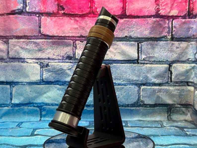 May include: A black and silver lightsaber with a brown accent ring, displayed on a black stand. The lightsaber has a textured grip and a silver hilt. The background is a colorful brick wall.
