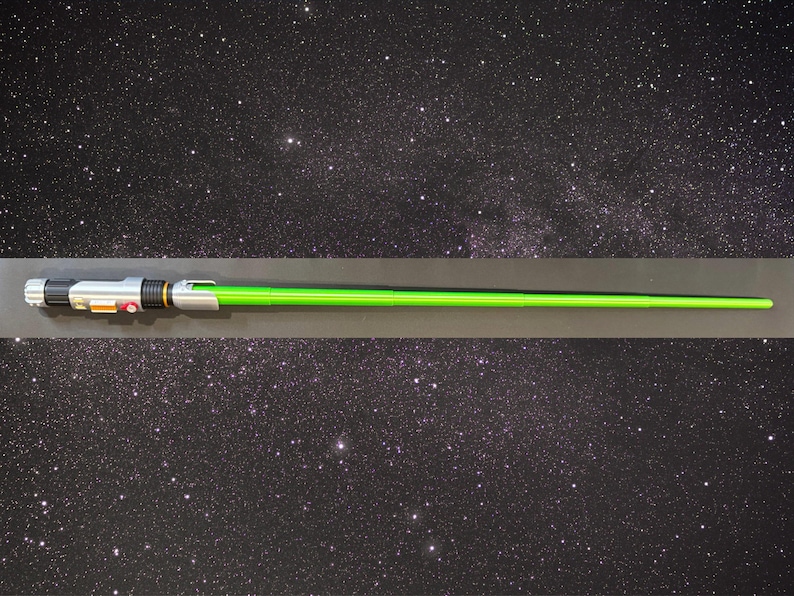 May include: A green lightsaber prop with a silver and black handle. The lightsaber is displayed against a starry background, suggesting a space or sci-fi theme. The handle has orange and red buttons.