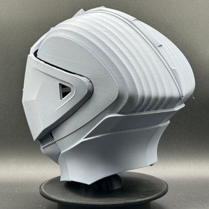 Puede incluir: Un casco blanco impreso en 3D con un dise&ntilde;o futurista. El casco tiene una visera y una superficie texturizada.