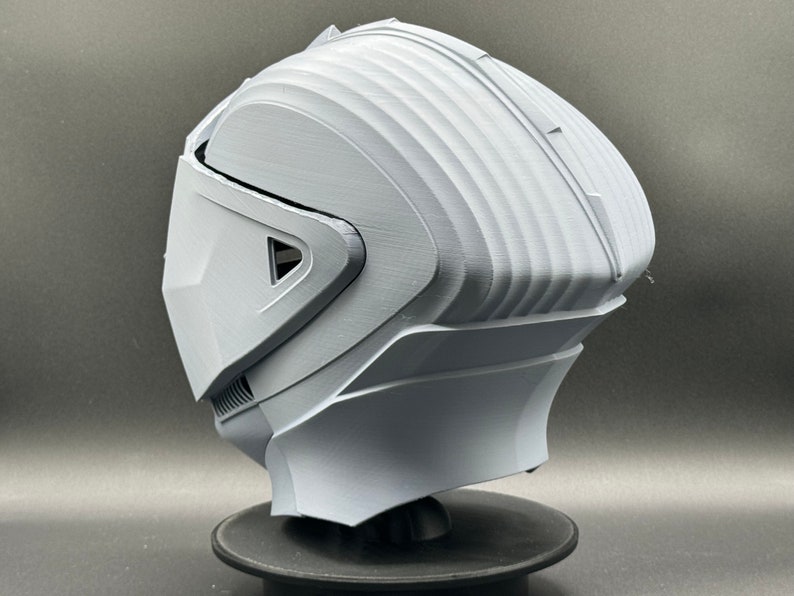 Puede incluir: Un casco blanco impreso en 3D con un dise&ntilde;o futurista. El casco tiene una visera y una superficie texturizada.