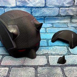 Puede incluir: Un casco negro texturizado con aberturas rojas para los ojos y cuernos peque&ntilde;os. Una pieza negra separada, posiblemente una placa frontal, y un cuerno peque&ntilde;o est&aacute;n junto al casco. Los art&iacute;culos est&aacute;n sobre una superficie con patr&oacute;n de ladrillos.