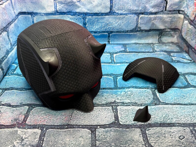 Puede incluir: Un casco negro texturizado con aberturas rojas para los ojos y cuernos peque&ntilde;os. Una pieza negra separada, posiblemente una placa frontal, y un cuerno peque&ntilde;o est&aacute;n junto al casco. Los art&iacute;culos est&aacute;n sobre una superficie con patr&oacute;n de ladrillos.