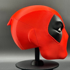 Puede incluir: Una m&aacute;scara de Deadpool roja y negra en un soporte negro. La m&aacute;scara tiene una superficie texturizada y un dise&ntilde;o detallado.