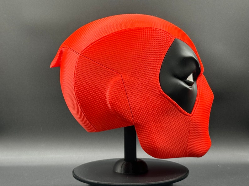 Puede incluir: Una m&aacute;scara de Deadpool roja y negra en un soporte negro. La m&aacute;scara tiene una superficie texturizada y un dise&ntilde;o detallado.