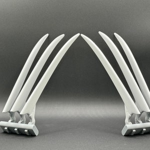 May include: A pair of gray plastic Wolverine claws, each with three claws attached to a base. The claws are curved and pointed, and the bases are rectangular.