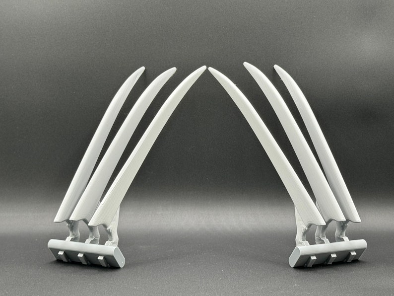 May include: A pair of gray plastic Wolverine claws, each with three claws attached to a base. The claws are curved and pointed, and the bases are rectangular.