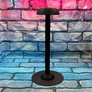 May include: A black hat stand with a round base and a tall, slender pole. The top of the stand has a curved, hat-shaped holder. The stand is made of metal and is designed to display hats.