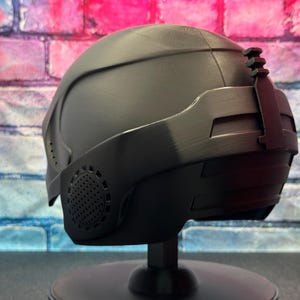 May include: A matte black helmet with a visor and a textured surface. The helmet has a series of vents and a small attachment on the top. It is displayed on a black stand against a brick wall.