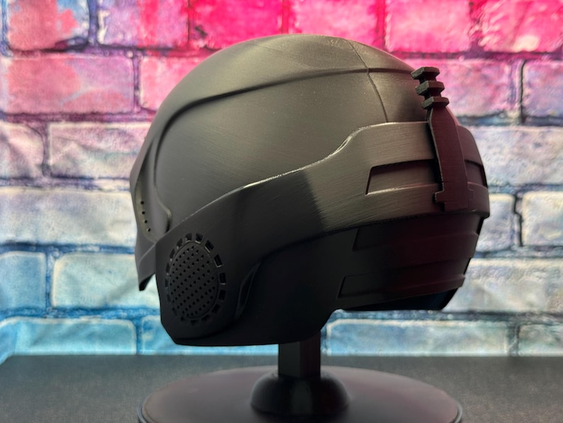 May include: A matte black helmet with a visor and a textured surface. The helmet has a series of vents and a small attachment on the top. It is displayed on a black stand against a brick wall.