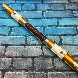 Puede incluir: Un sable de luz dorado y blanco con empuñadura marrón. El sable de luz tiene un acabado metálico con detalles plateados. El mango es marrón con una serie de líneas horizontales. El sable de luz mide unos 30 cm de largo.