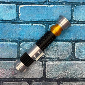 May include: A replica lightsaber with a silver, gold, and black design. The hilt has a black grip with circular indentations. The top and bottom are silver, with a gold section in the middle. The lightsaber is a prop.